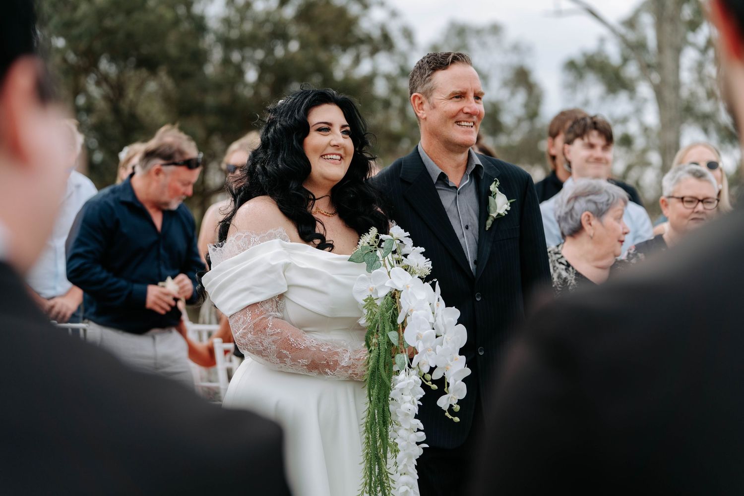 Wedding guests gather outdoors for a celebration with white floral bouquets and formal attire.