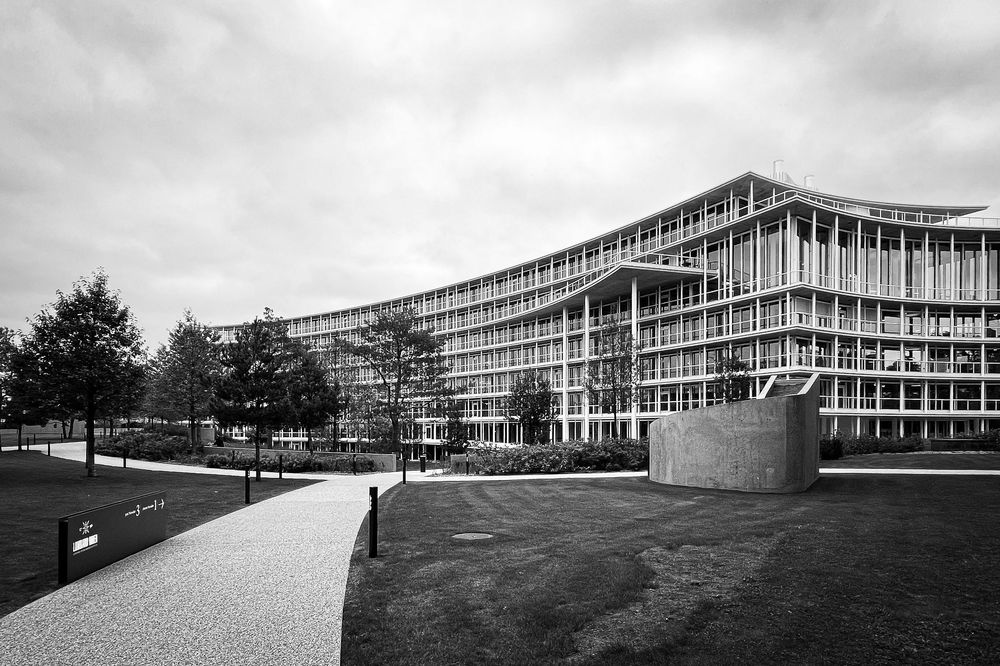 Exterior view of a modernist school building with glass curtain walls and horizontal lines under an overcast sky.