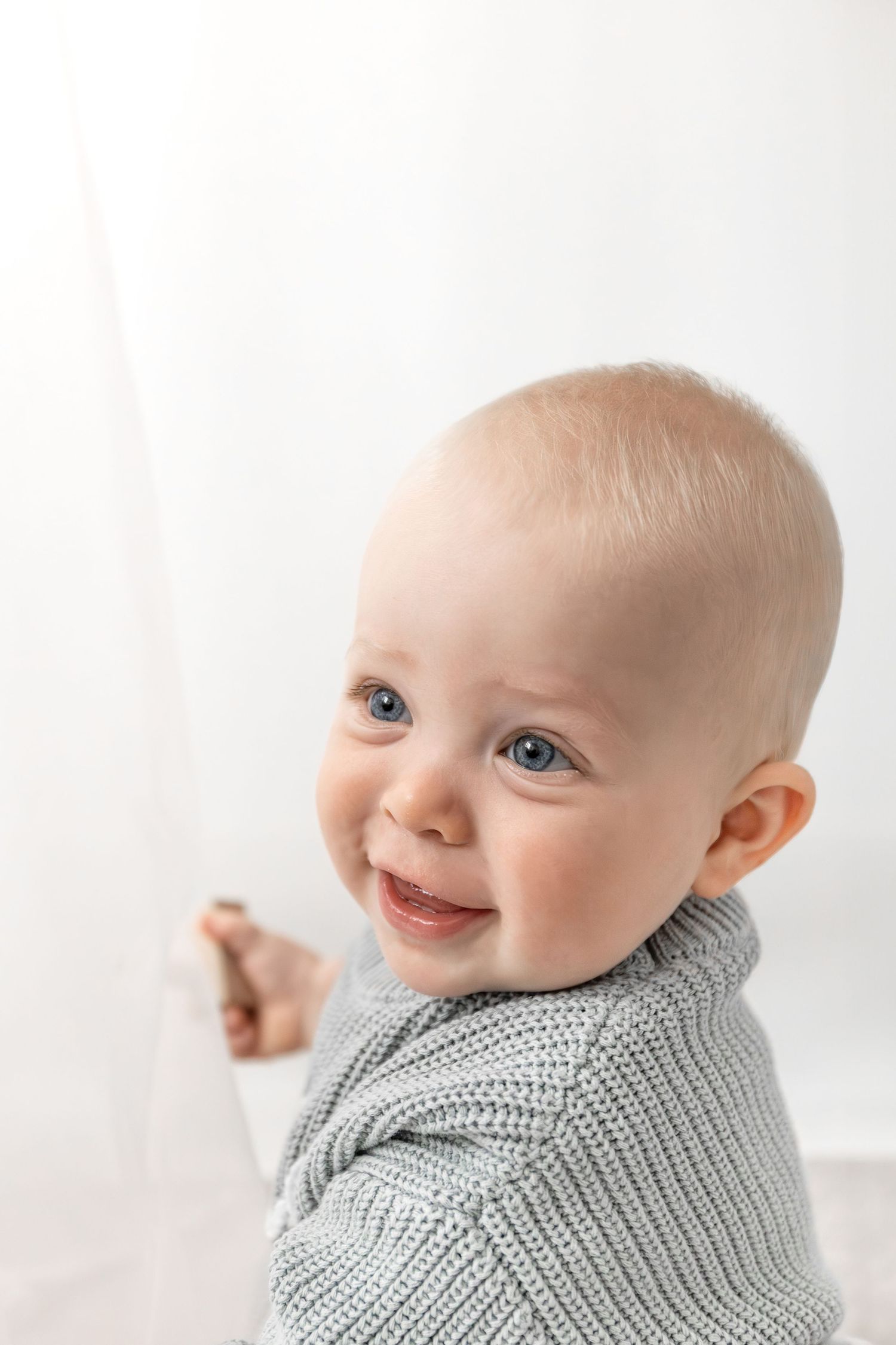 Baby wearing cozy blue knit jumper smiles sweetly in bright white studio setting, during birthday shoot in Nottingham