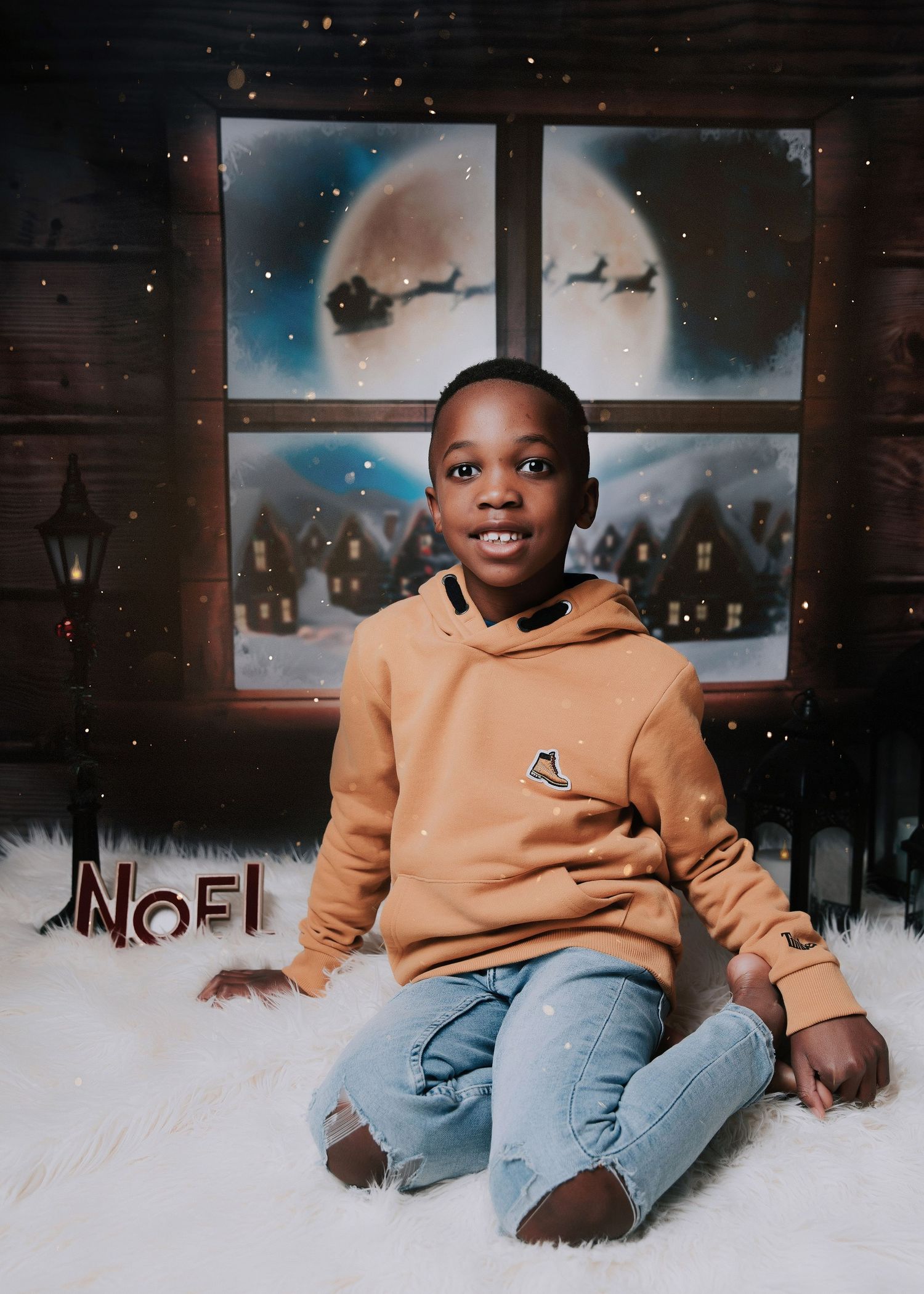 A child wearing a tan hoodie and jeans sits on white fluffy carpet in front of winter-themed window decorations.