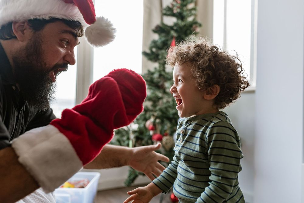 A person in a Santa hat interacts with a joyful curly-haired toddler in front of a Christmas tree.