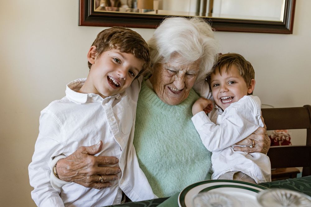 Three generations share joyful laughter while embracing on a couch in a sunlit living room.