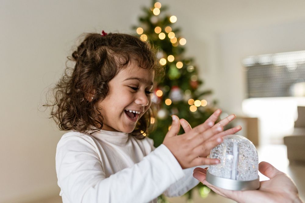 A young child in white clothing opens a gift box in front of a decorated Christmas tree.