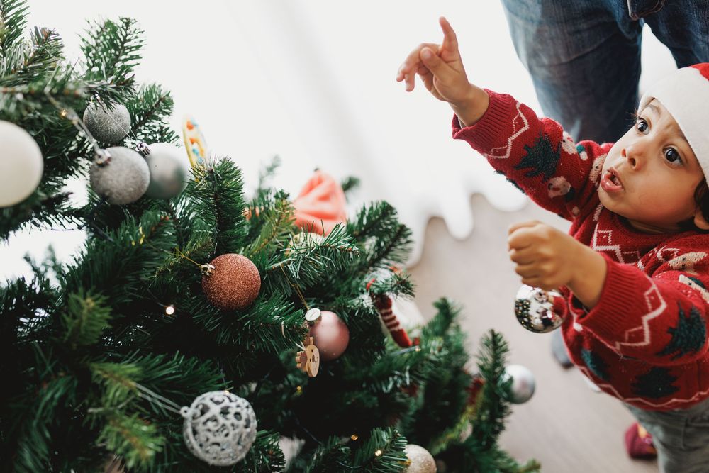A hand reaches up to place a silver ornament on a green Christmas tree decorated with festive holiday decorations.