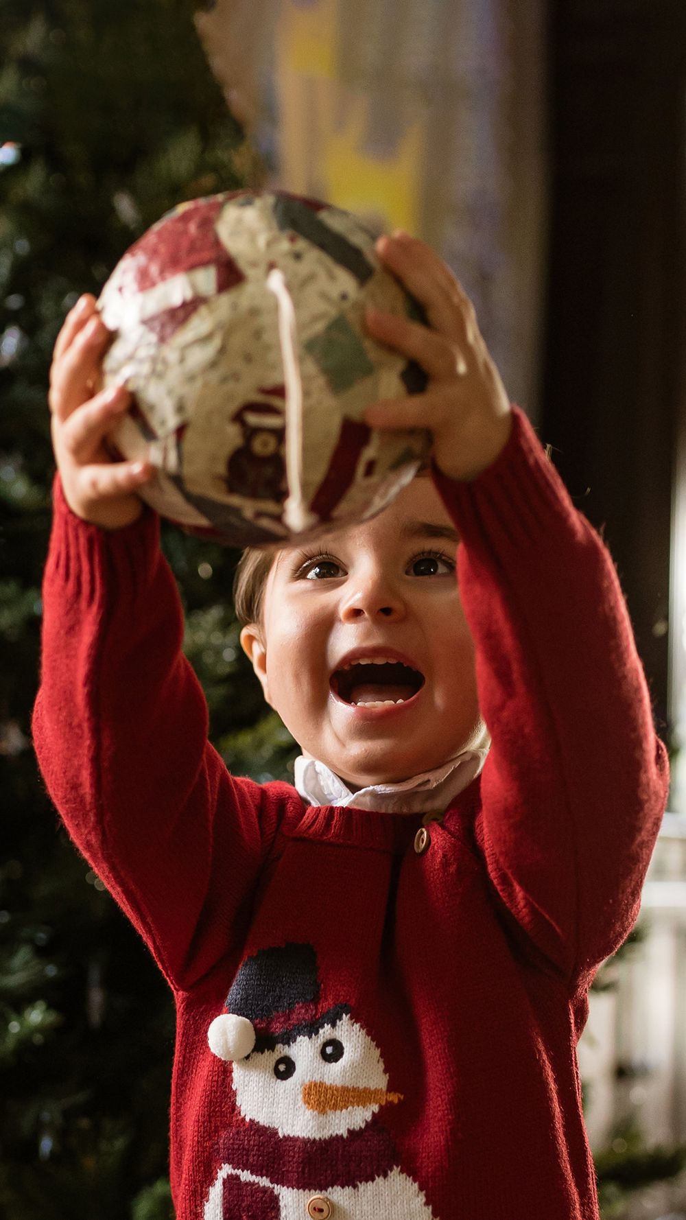 Child in red snowman sweater excitedly holds up a Christmas ornament with arms raised during holiday decorating.