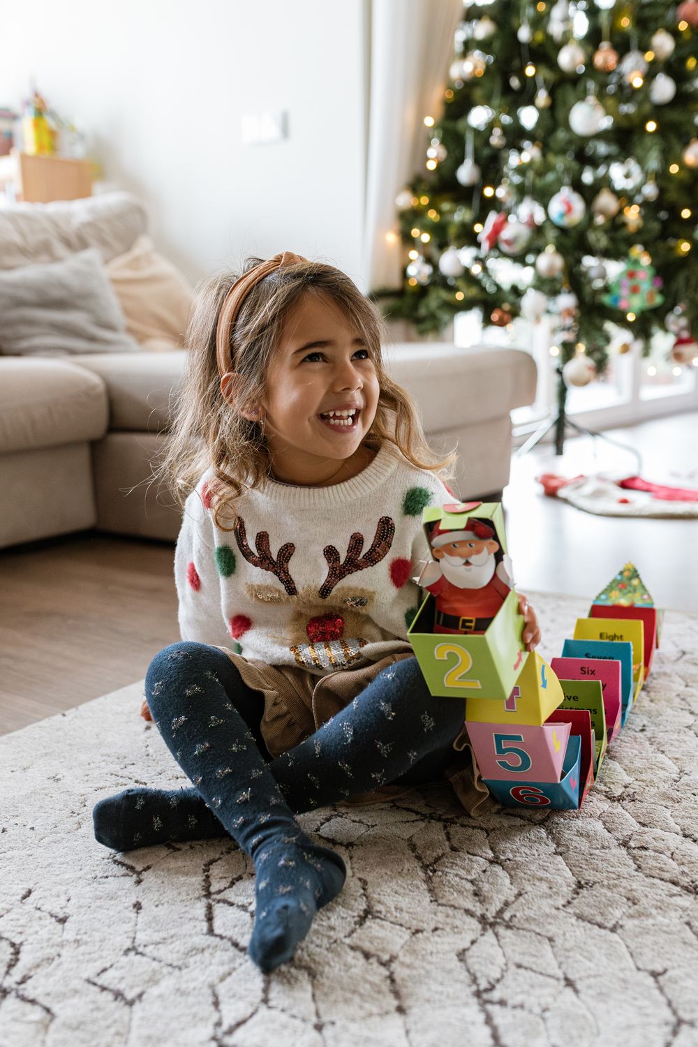 A child sits on a patterned rug playing with colorful numbered blocks in front of a decorated Christmas tree.