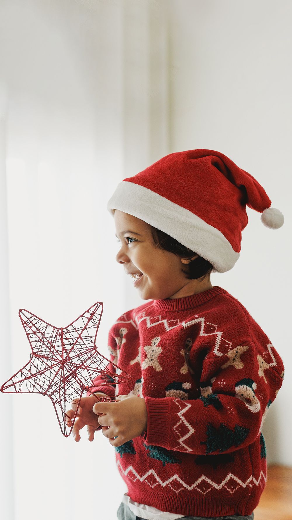 Child in red Christmas sweater and Santa hat holds decorative star while smiling during holiday season.