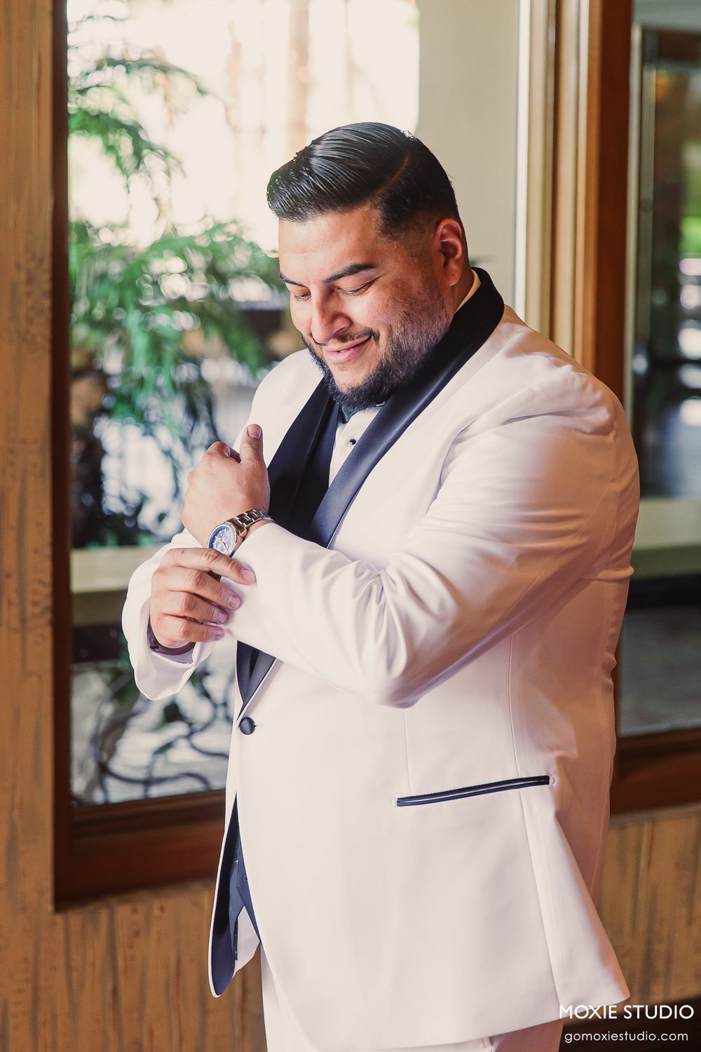 A groom in a white tuxedo jacket laughs while getting ready in a wood-paneled room with natural lighting.