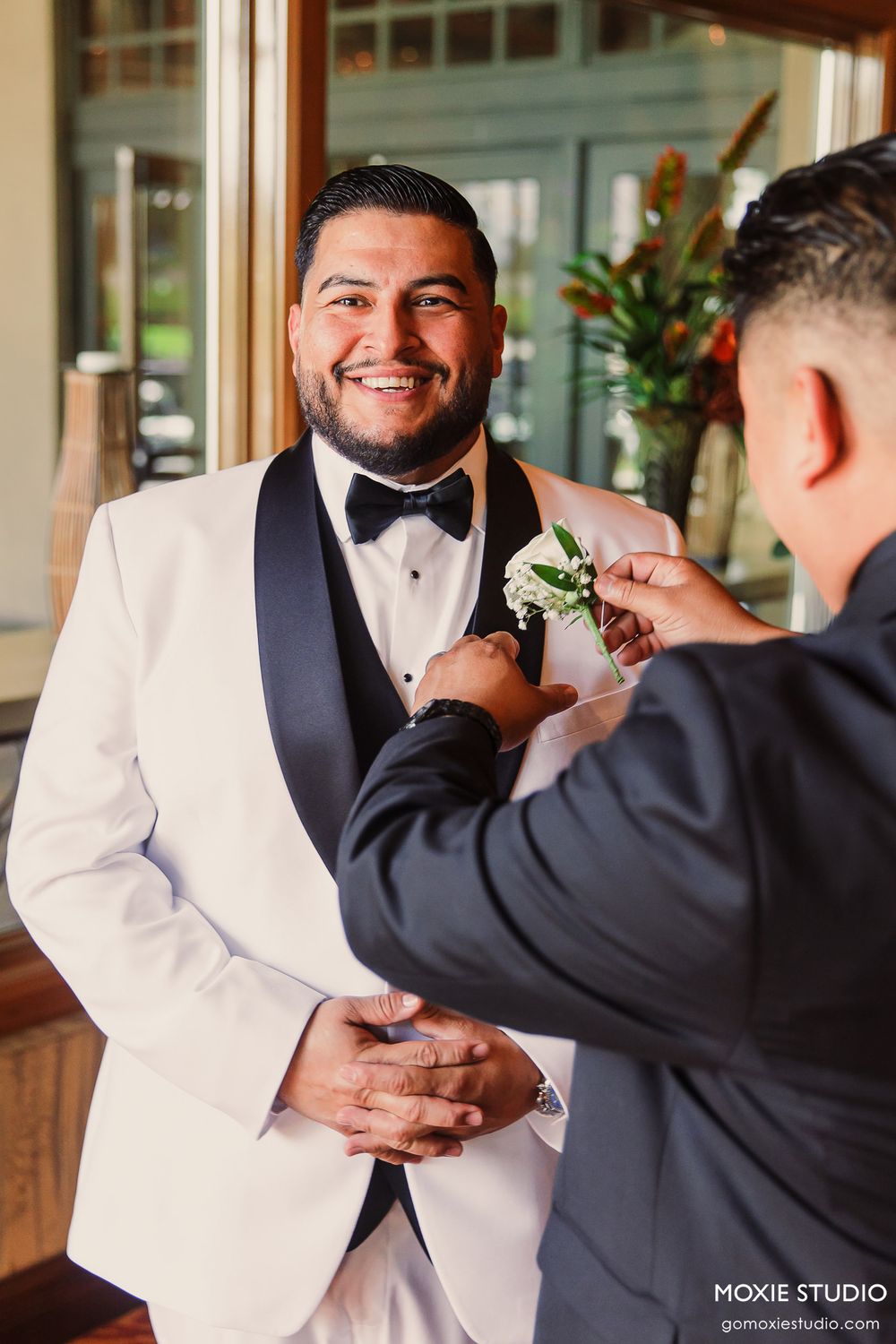 A groomsman in a white tuxedo jacket and black bow tie smiles while having a boutonniere pinned on by another person.