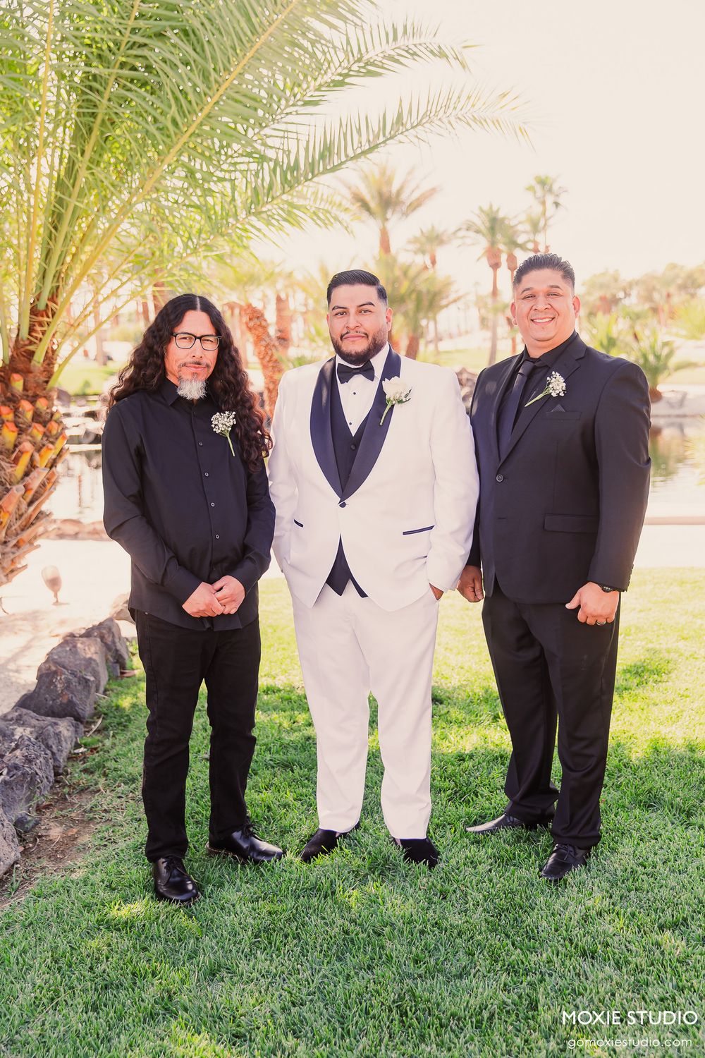 A group in formal attire poses together on grass with palm trees in the background at an outdoor event.