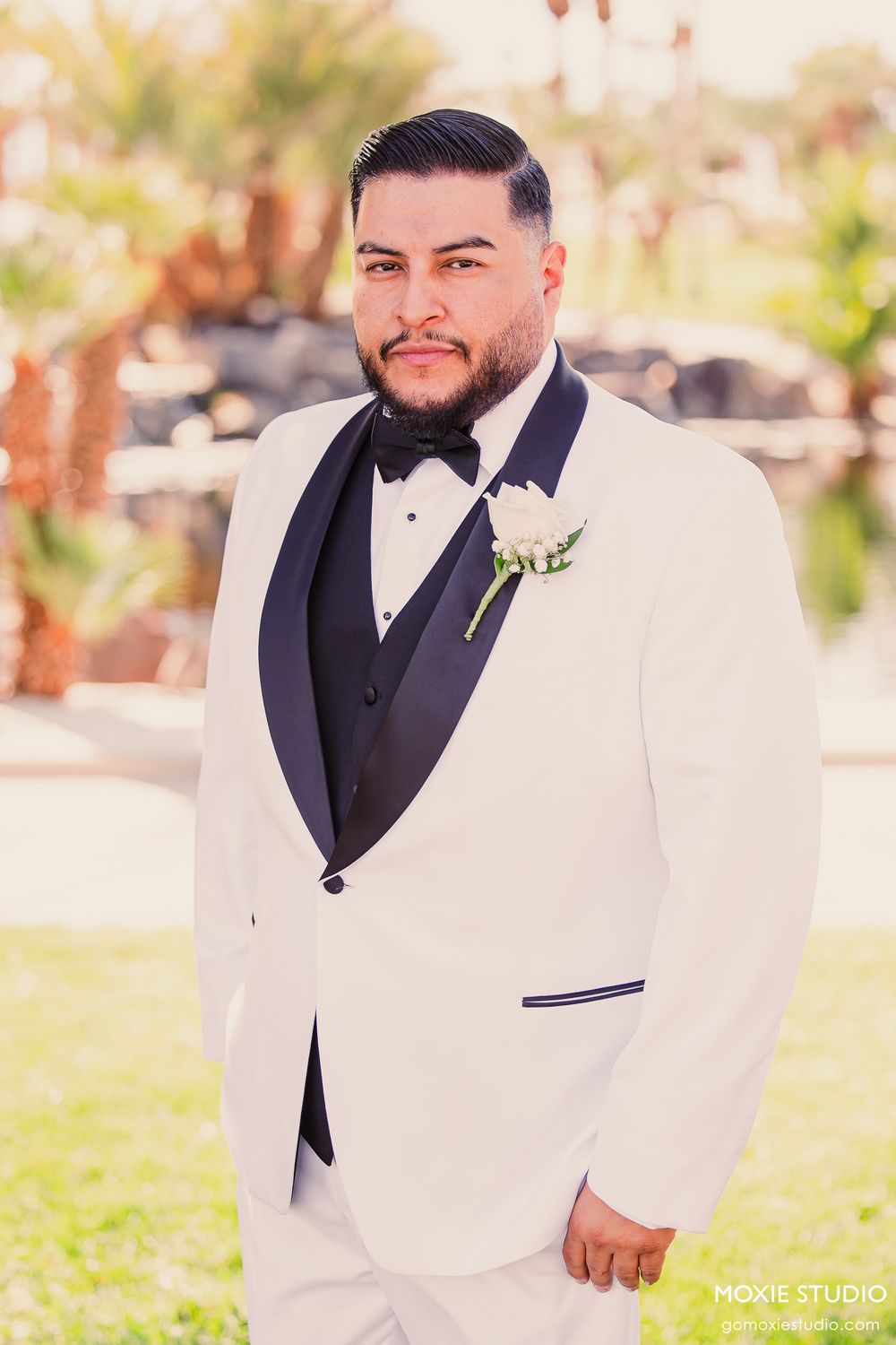 A groom in a white tuxedo jacket with black lapels and bow tie poses outdoors for formal wedding portraits.