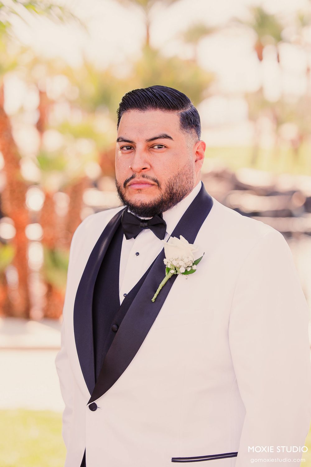 Groom in a classic white tuxedo jacket and black bow tie poses outdoors for wedding photos.