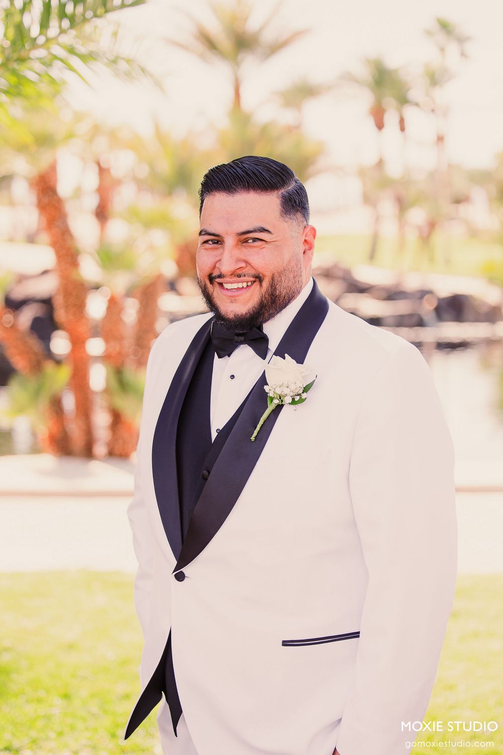 A groom in a white tuxedo jacket and black bow tie smiles outdoors during a wedding photoshoot.