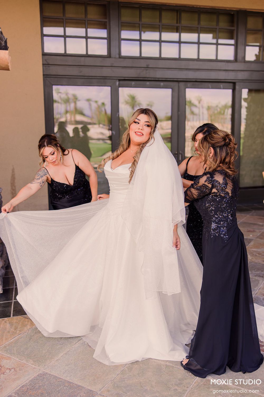 A bride in a flowing white wedding gown laughs joyfully while two women in formal dresses help arrange her dress and veil.