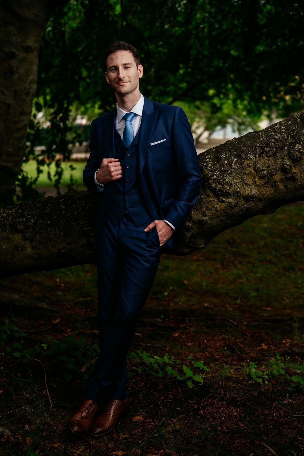 A person in a navy blue suit poses casually against a tree in a wooded outdoor setting.