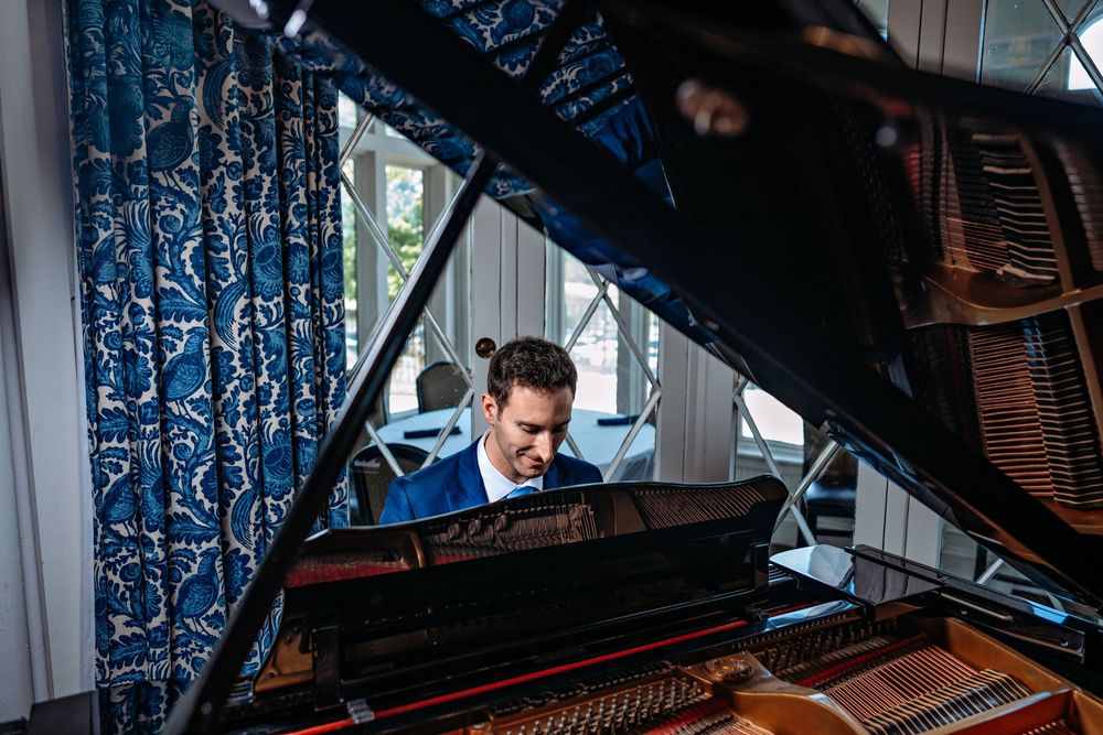 Pianist performing at a grand piano by a window with blue curtains in natural light.