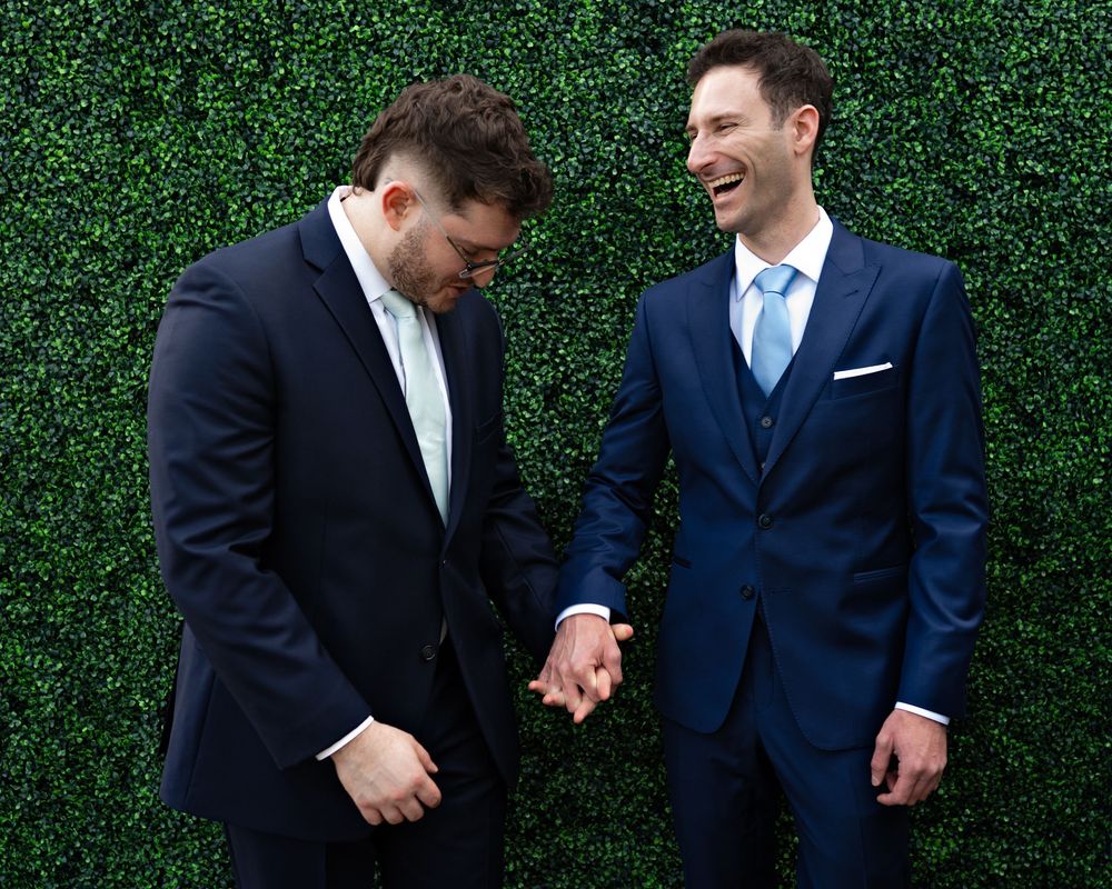 Two people in navy suits share a candid moment against a lush green hedge wall backdrop.