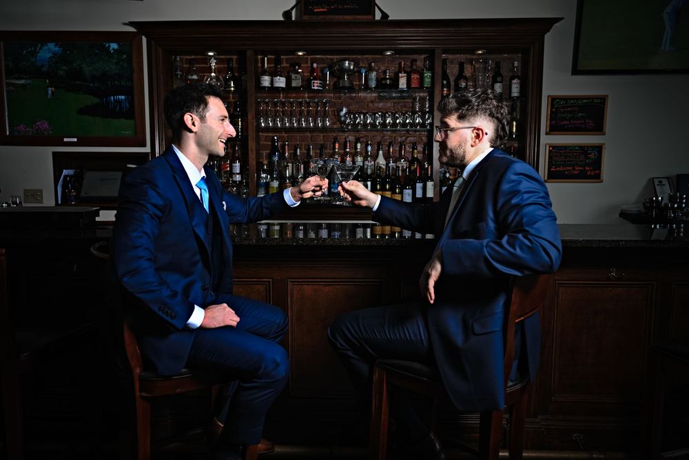 Two men in blue suits sit at a bar counter having a conversation in a dimly lit setting.
