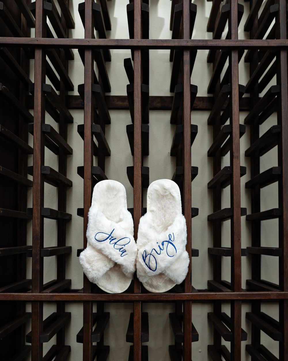 White bridal shoes with 'I do' written in blue script lettering rest on dark wooden wine rack slats.