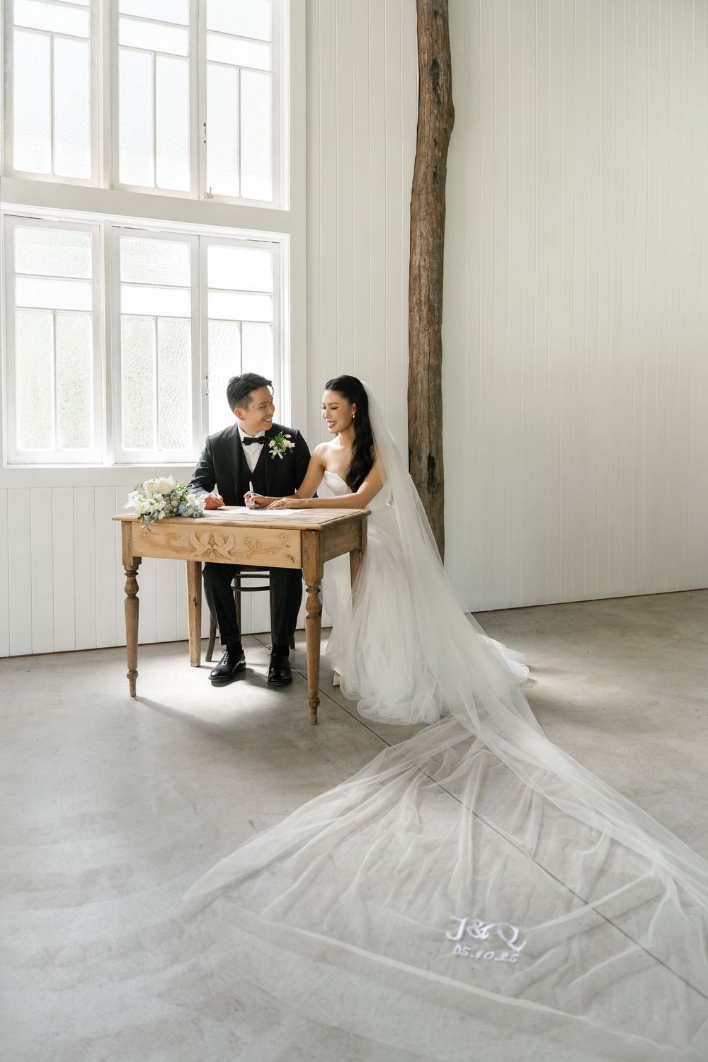 A bride and groom sign documents at a rustic wooden table in a bright minimalist room.