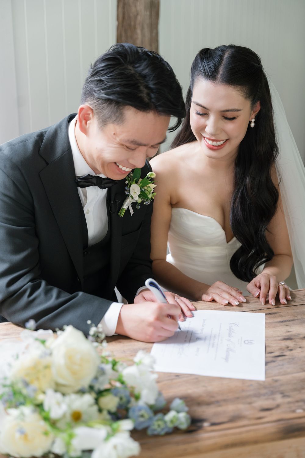 Couple signing marriage documents at rustic wooden table with delicate floral centerpiece.