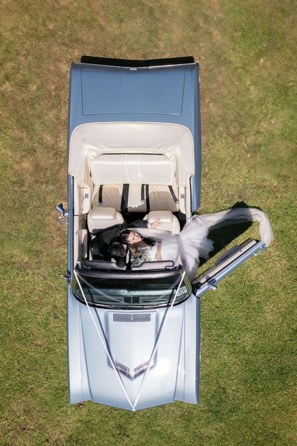 Aerial view of newlyweds posing in classic light blue vintage car on grassy lawn.