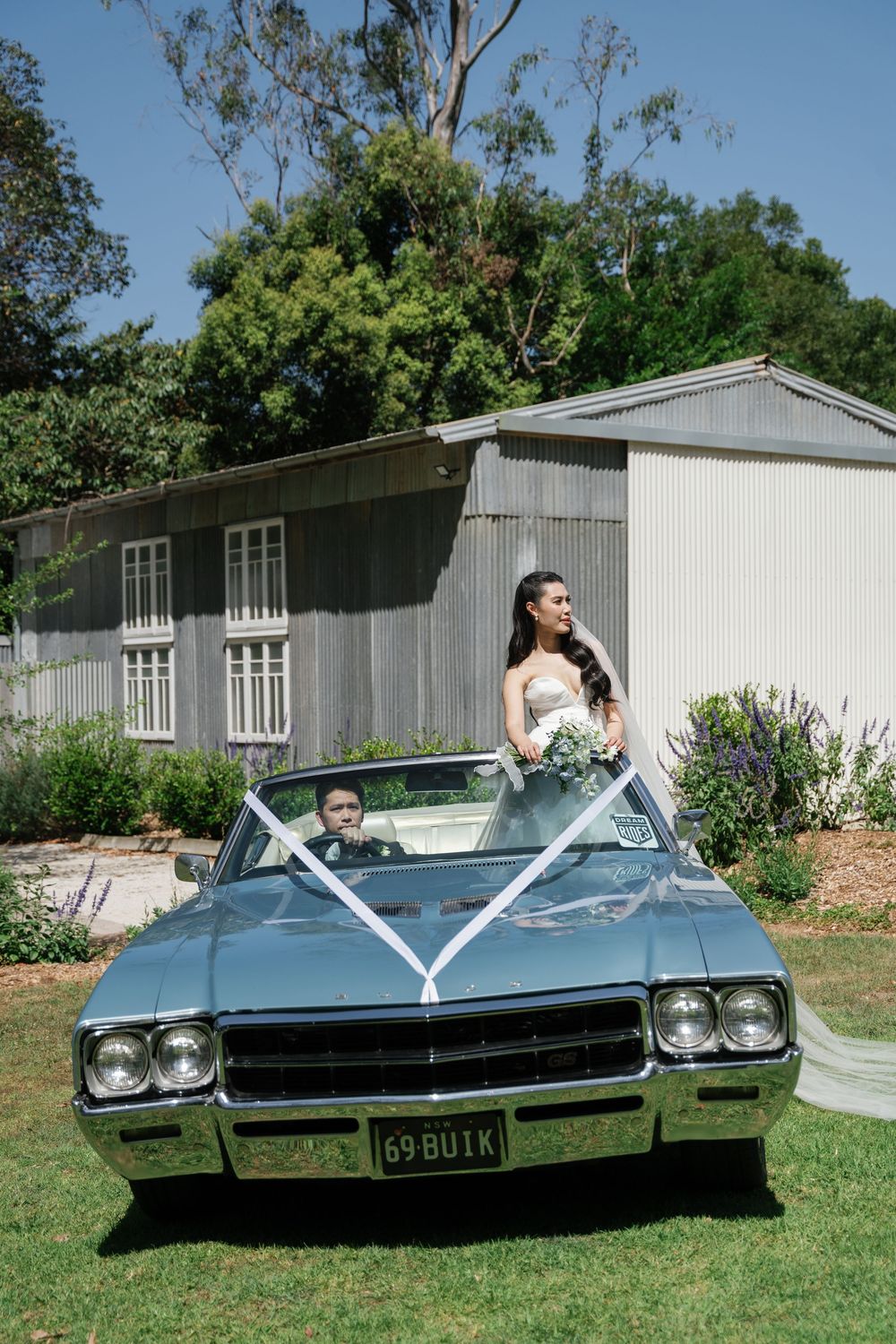 Classic blue Buick convertible parked on grass with white buildings in background.