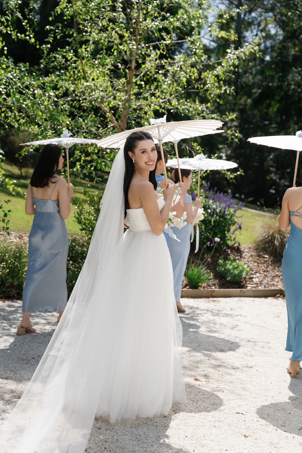 Bride in flowing white gown and veil walks along garden path with bridesmaids in light blue dresses on sunny day.
