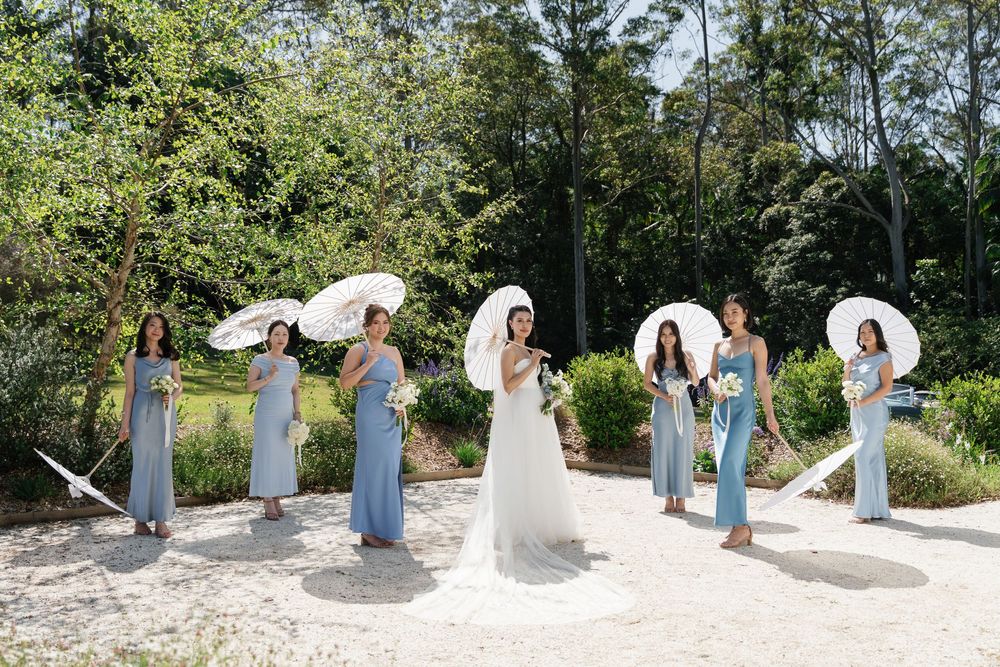 A bridal party poses with white parasols on a sunny garden path wearing light blue bridesmaid dresses.