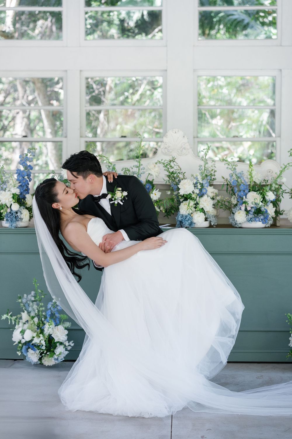 Romantic wedding couple in a dip pose with blue and white floral arrangements by a window.