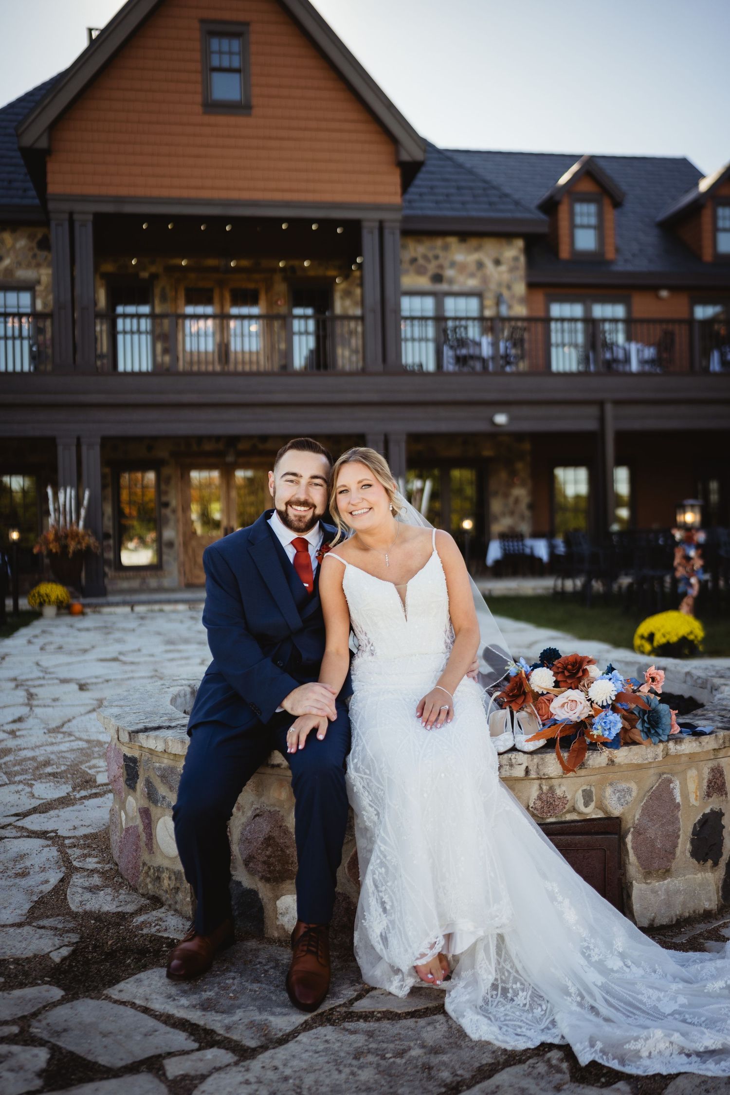 A couple sits together on a rustic stone wall outside a brown wooden lodge during their wedding celebration.