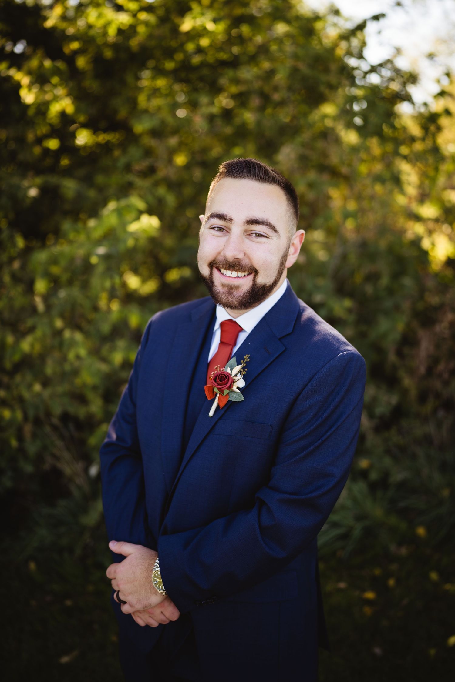 A groom in a navy blue suit and red tie poses outdoors during golden hour at a wedding photoshoot.