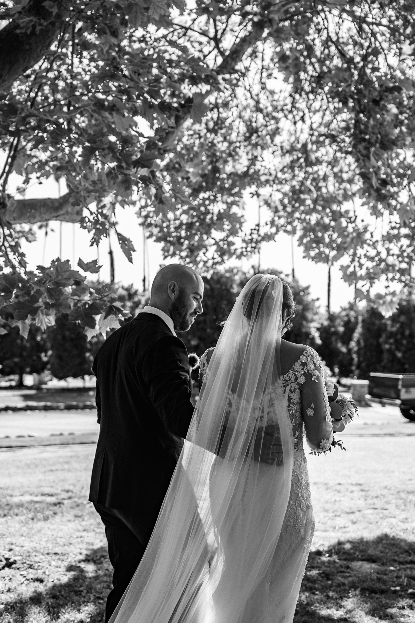 Black and white photo of couple walking outdoors with flowing bridal veil.