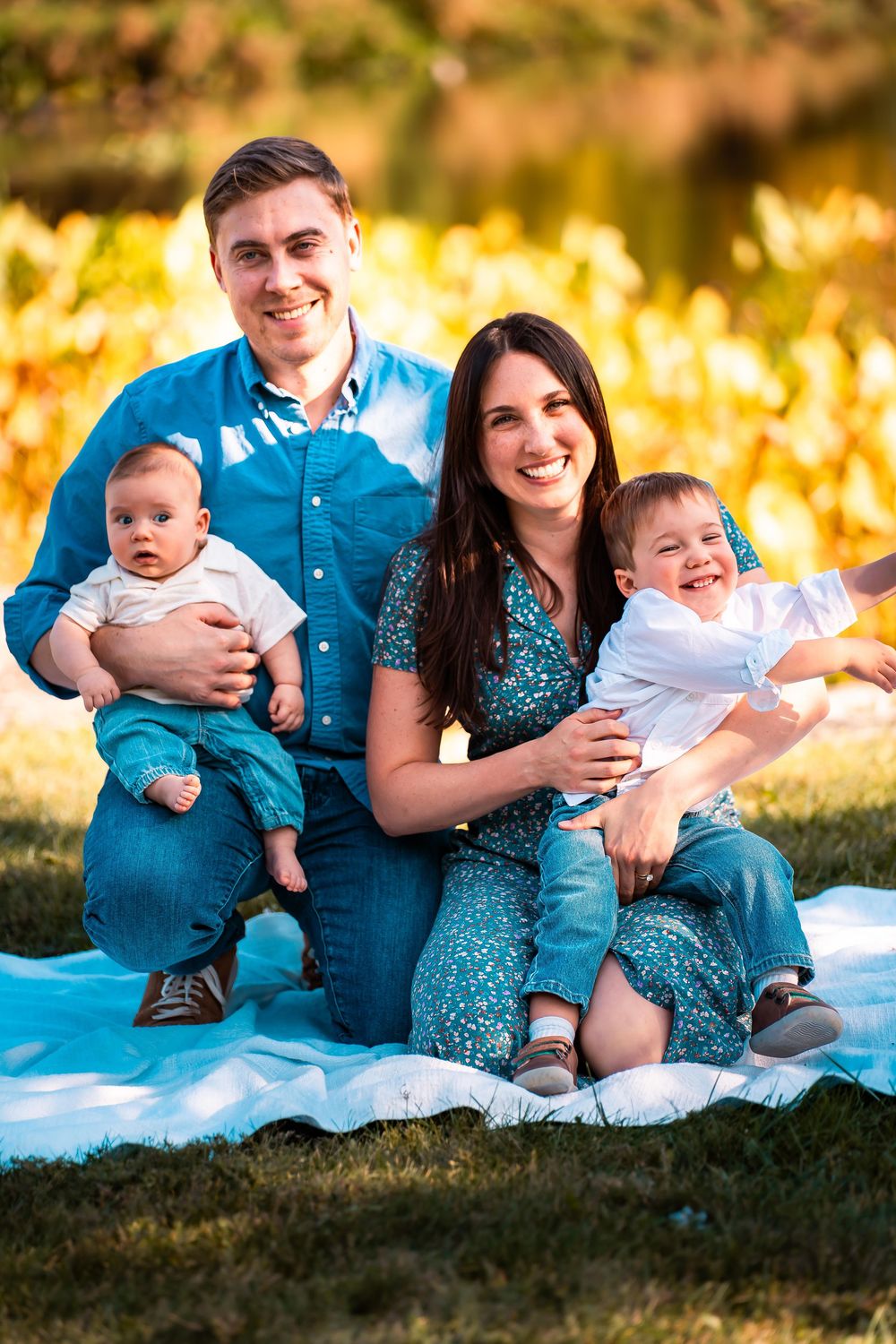 Family sitting on blue blanket outdoors with golden sunset lighting in background.