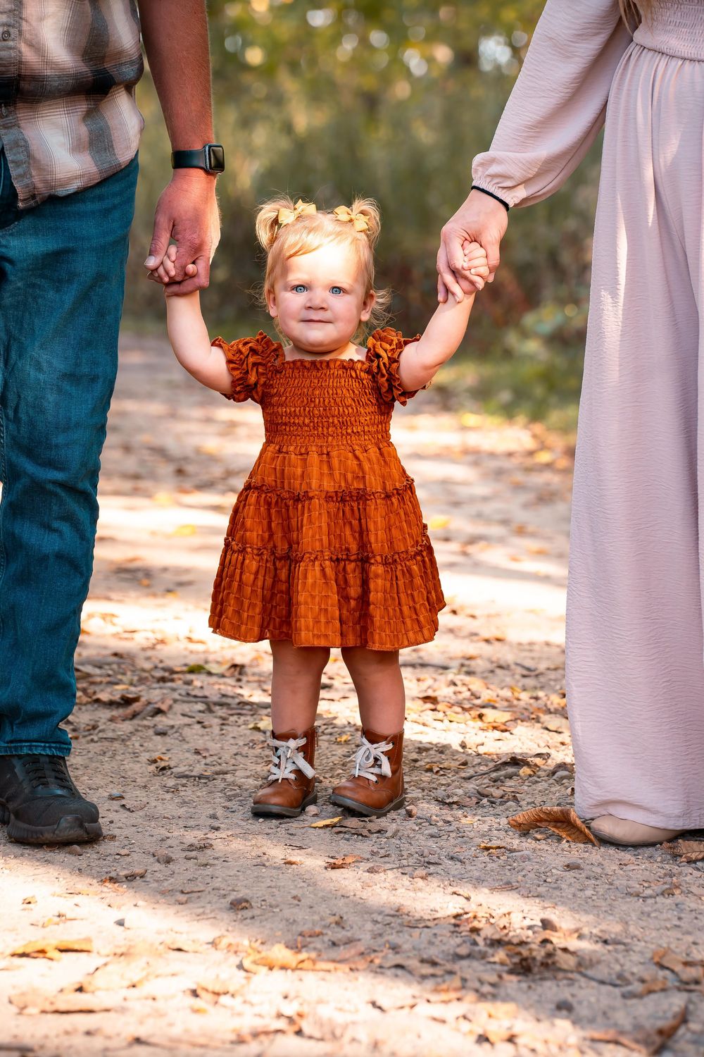 Close-up shots of toddler in rust-colored dress holding parents' hands on sandy path.