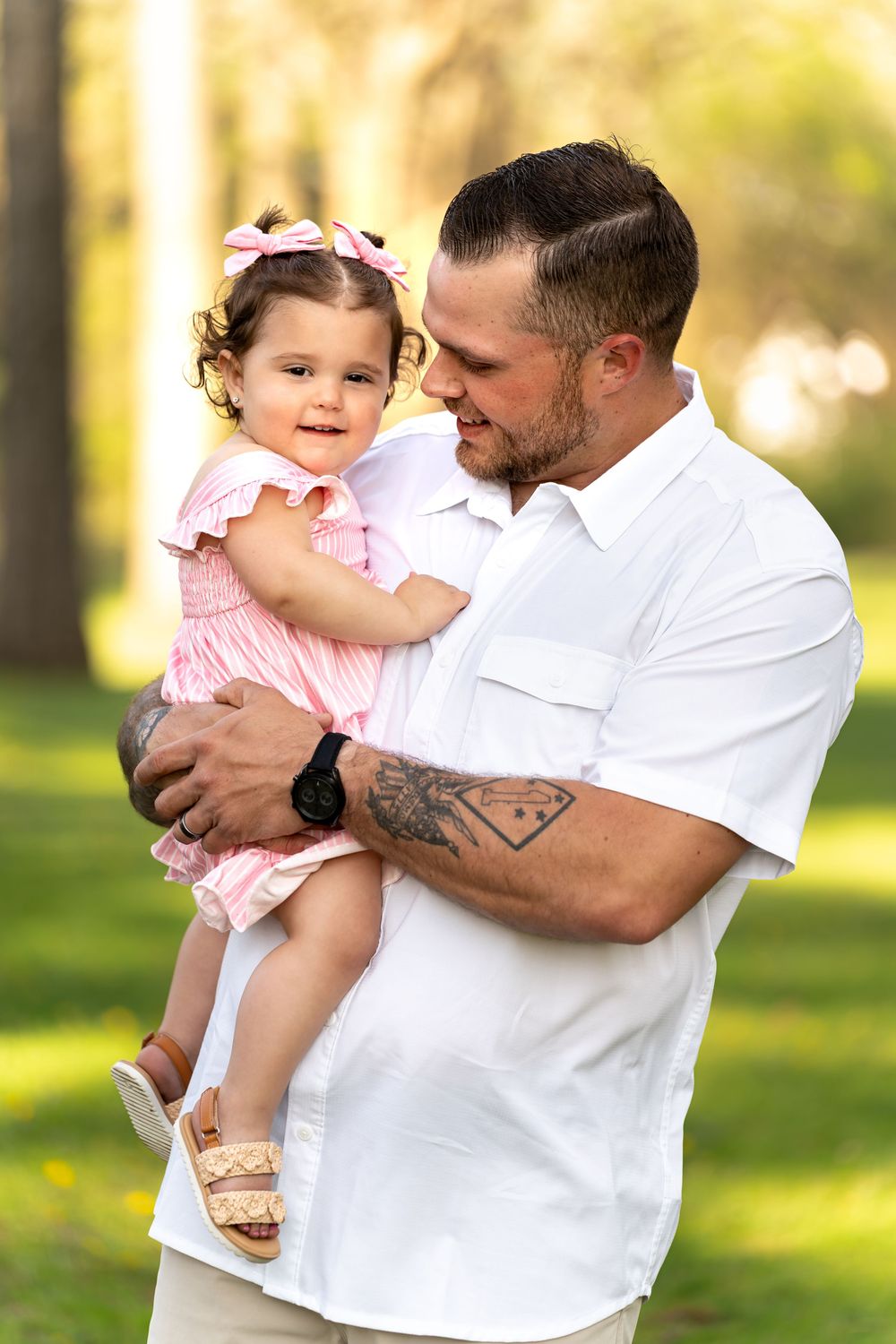 A tattooed father in a white shirt holds his smiling toddler daughter in a pink dress outdoors.