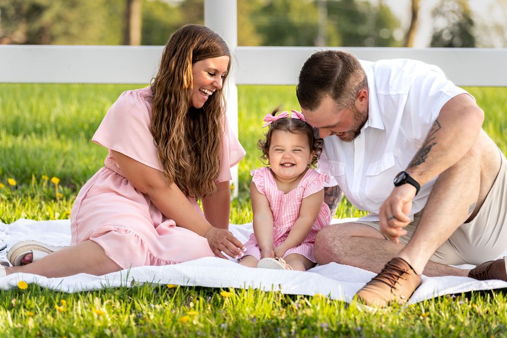 Happy family sitting on blanket in sunny green park, parents smiling at laughing toddler girl in pink dress.