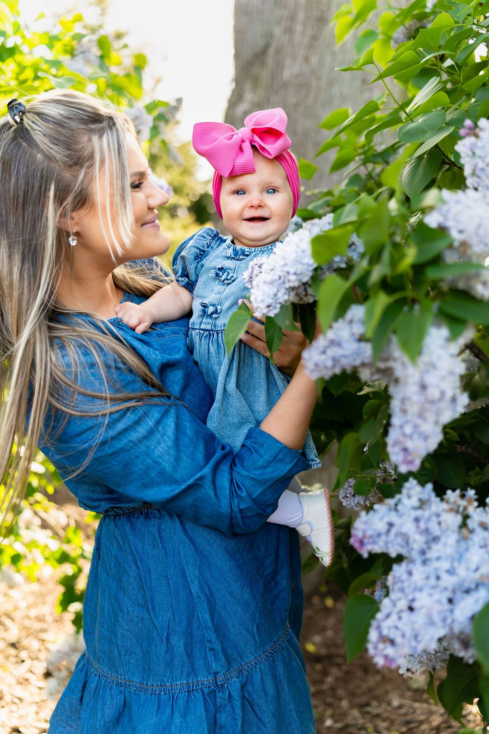 Mother in blue dress holds smiling baby girl with pink bow near blooming lilac bushes outdoors.