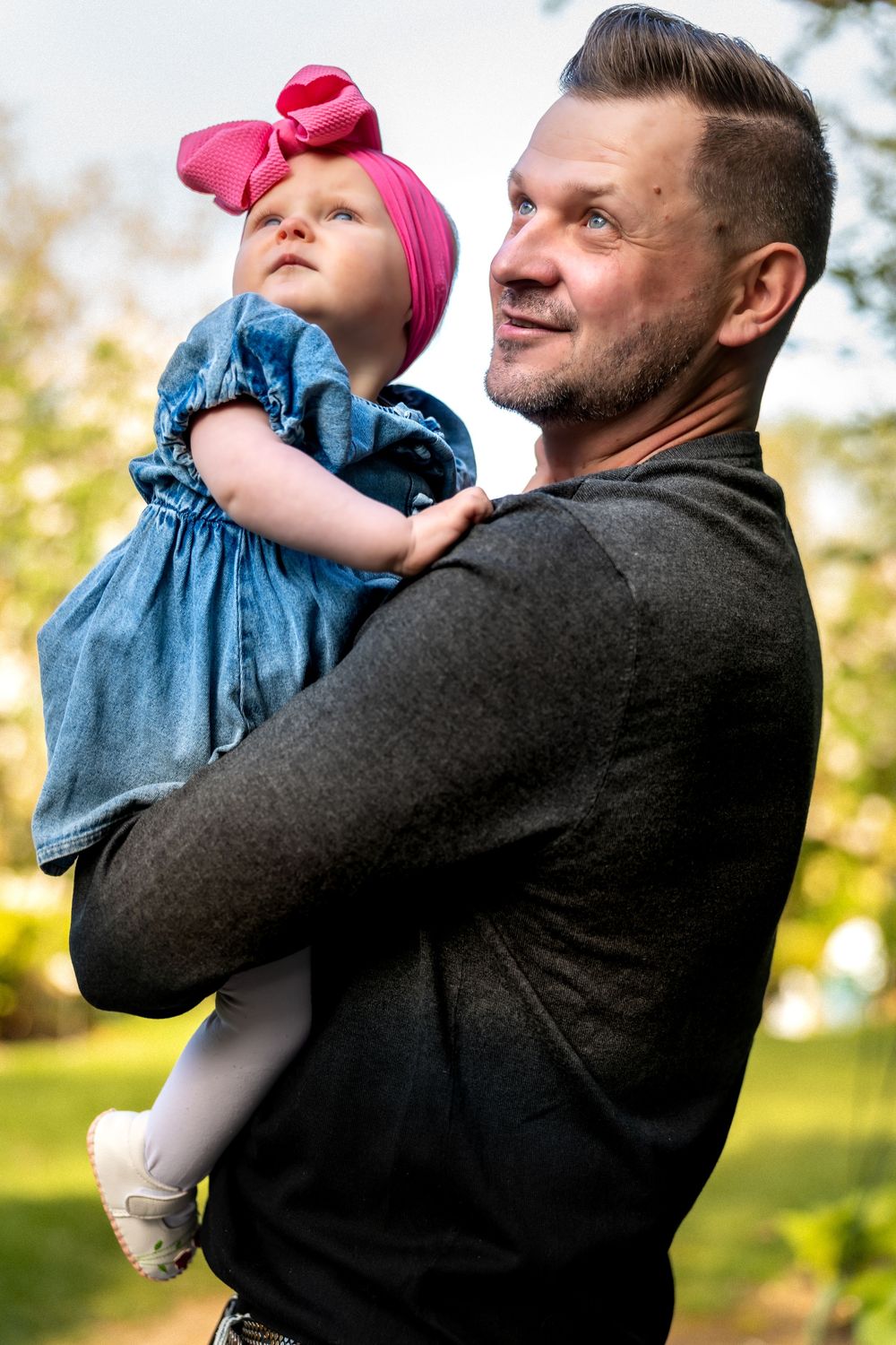 A father holds his baby daughter in a pink bow headband and denim dress outdoors in a green park setting.