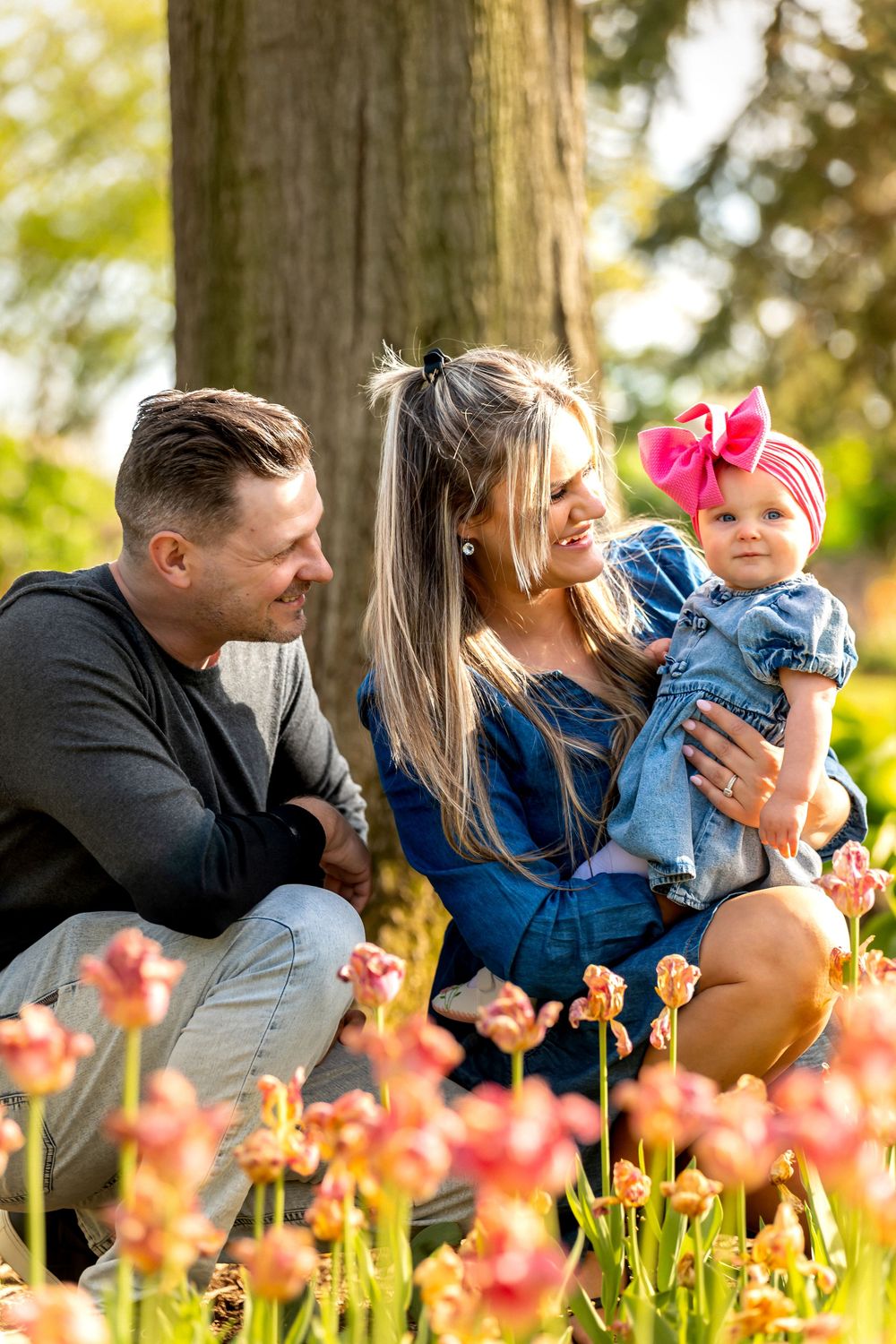 A happy family with a baby girl wearing a pink bow poses among blooming tulips in a sunny park.