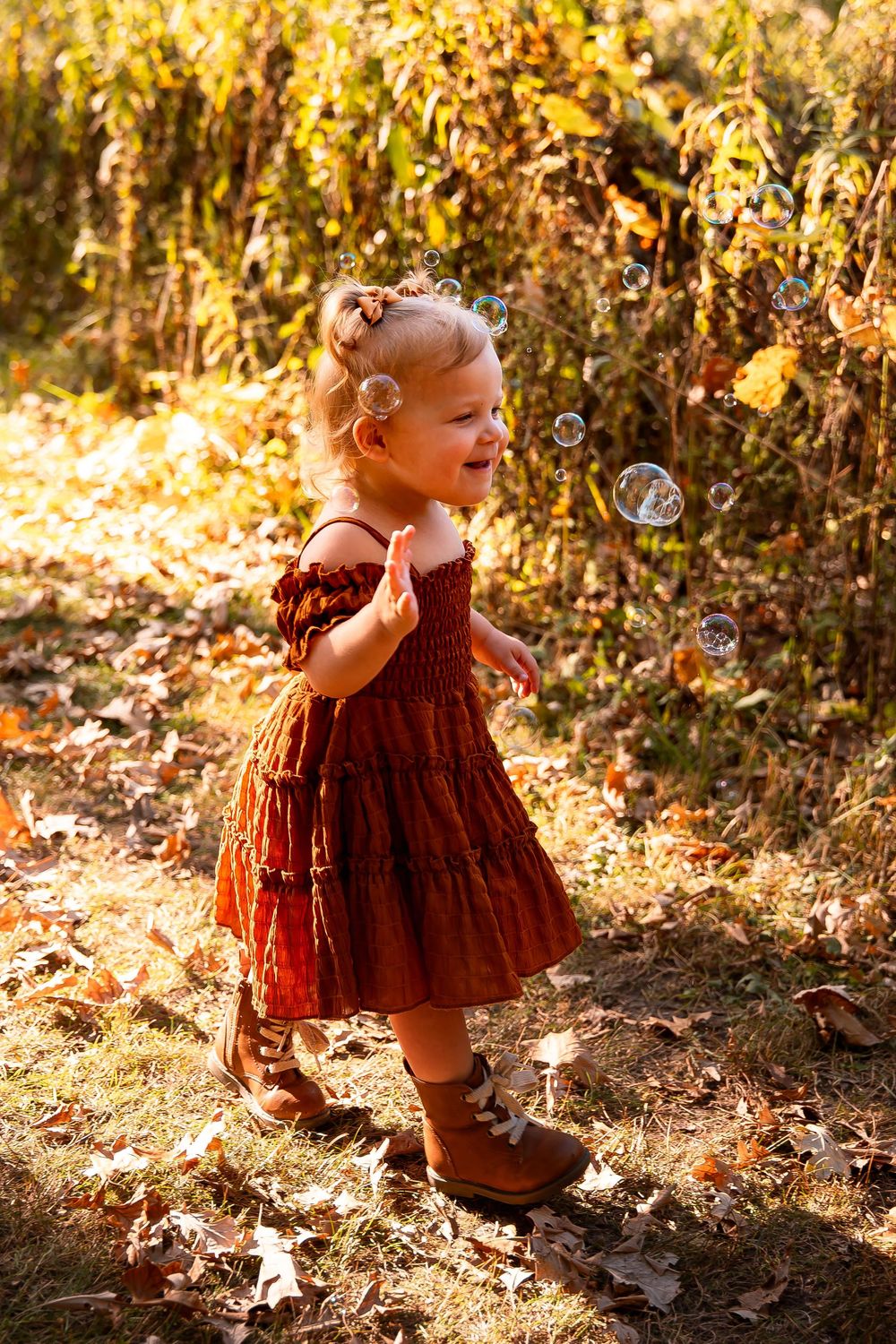 Toddler in rust-colored dress plays in fallen autumn leaves during golden hour.