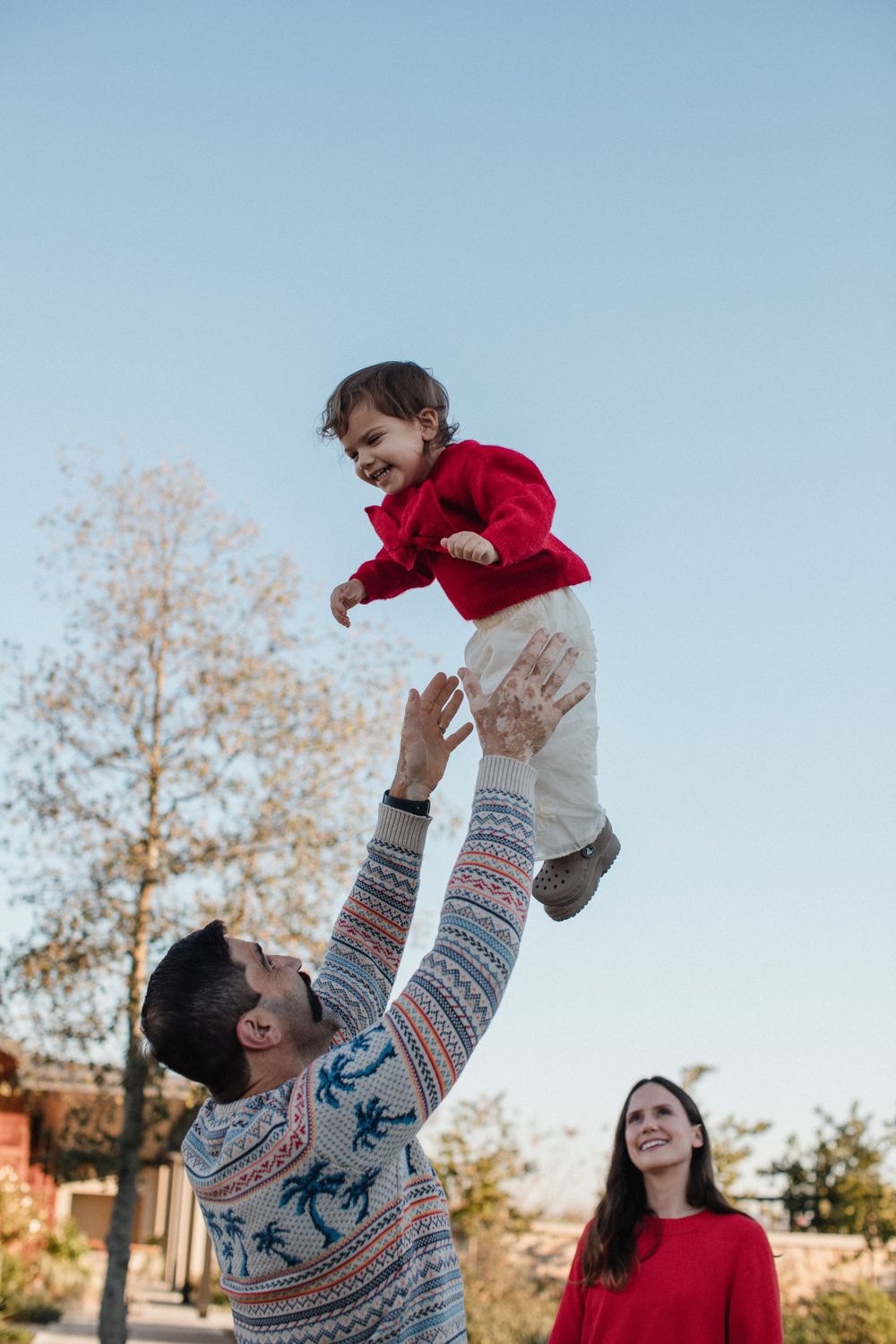 A family plays together outdoors on a clear autumn day, with a child being lifted up high in the air.