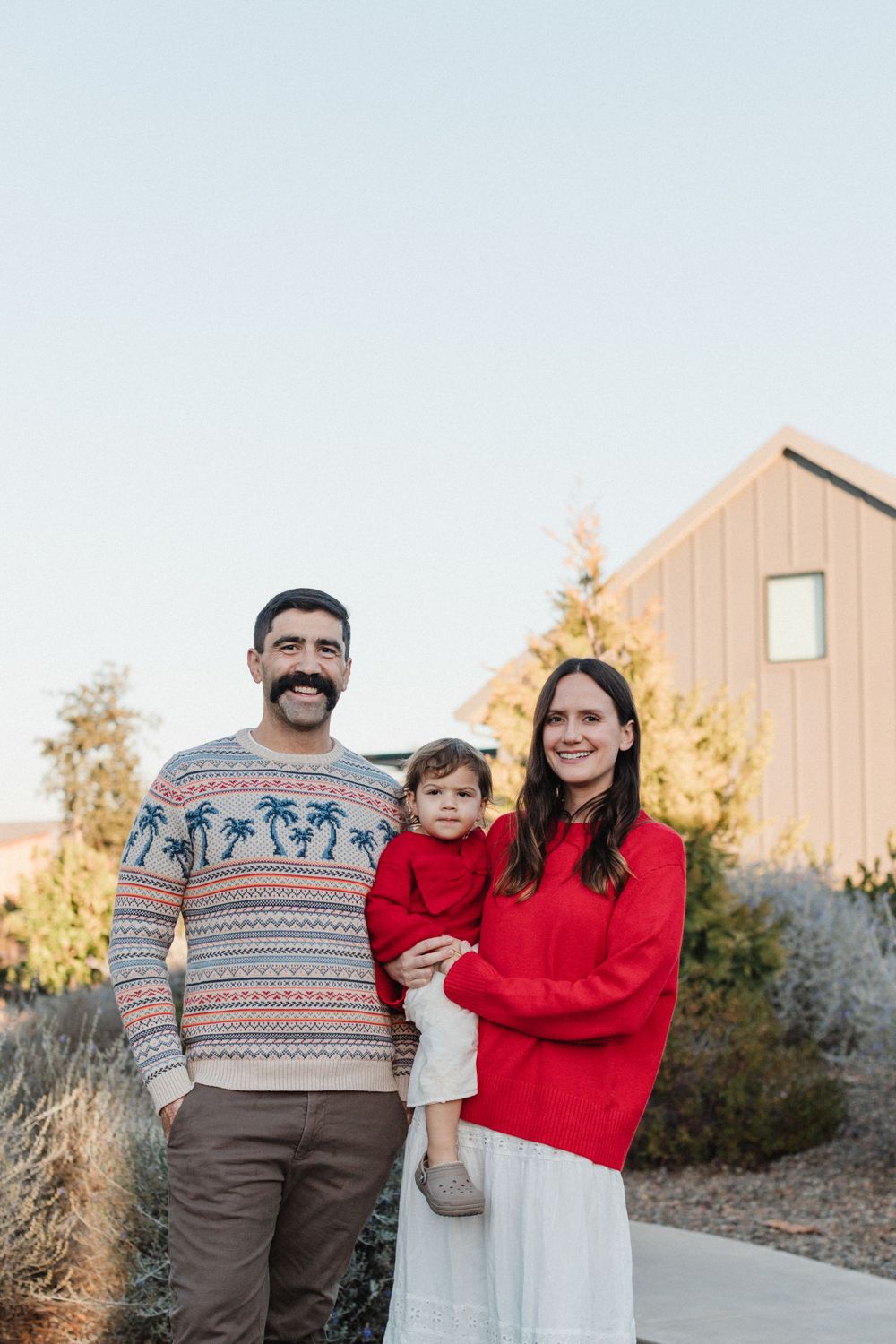 A family wearing festive sweaters stands outdoors in front of a wooden barn on a sunny autumn day.