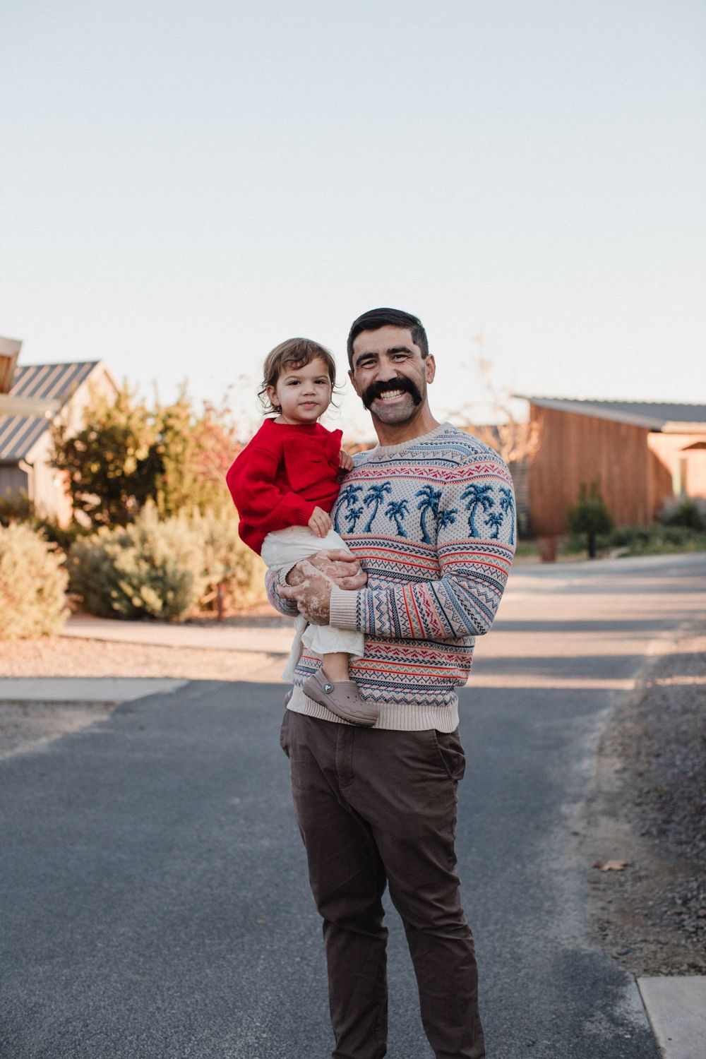 Standing in a residential street at sunset, someone wears a patterned winter sweater while holding a small child in a red coat.