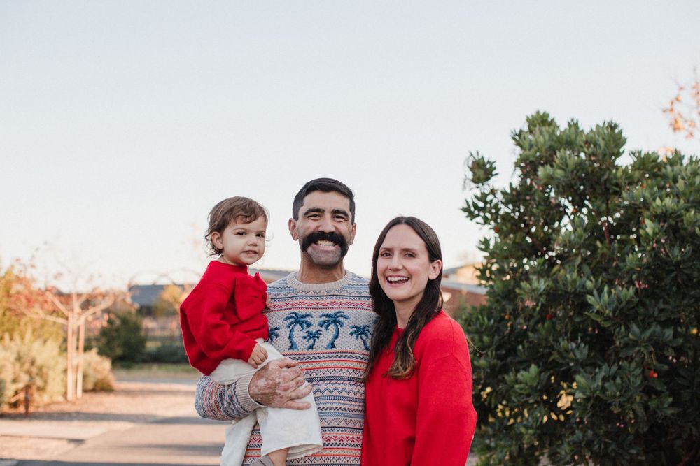 Three people wearing red and white clothing standing together outdoors at sunset near green foliage.