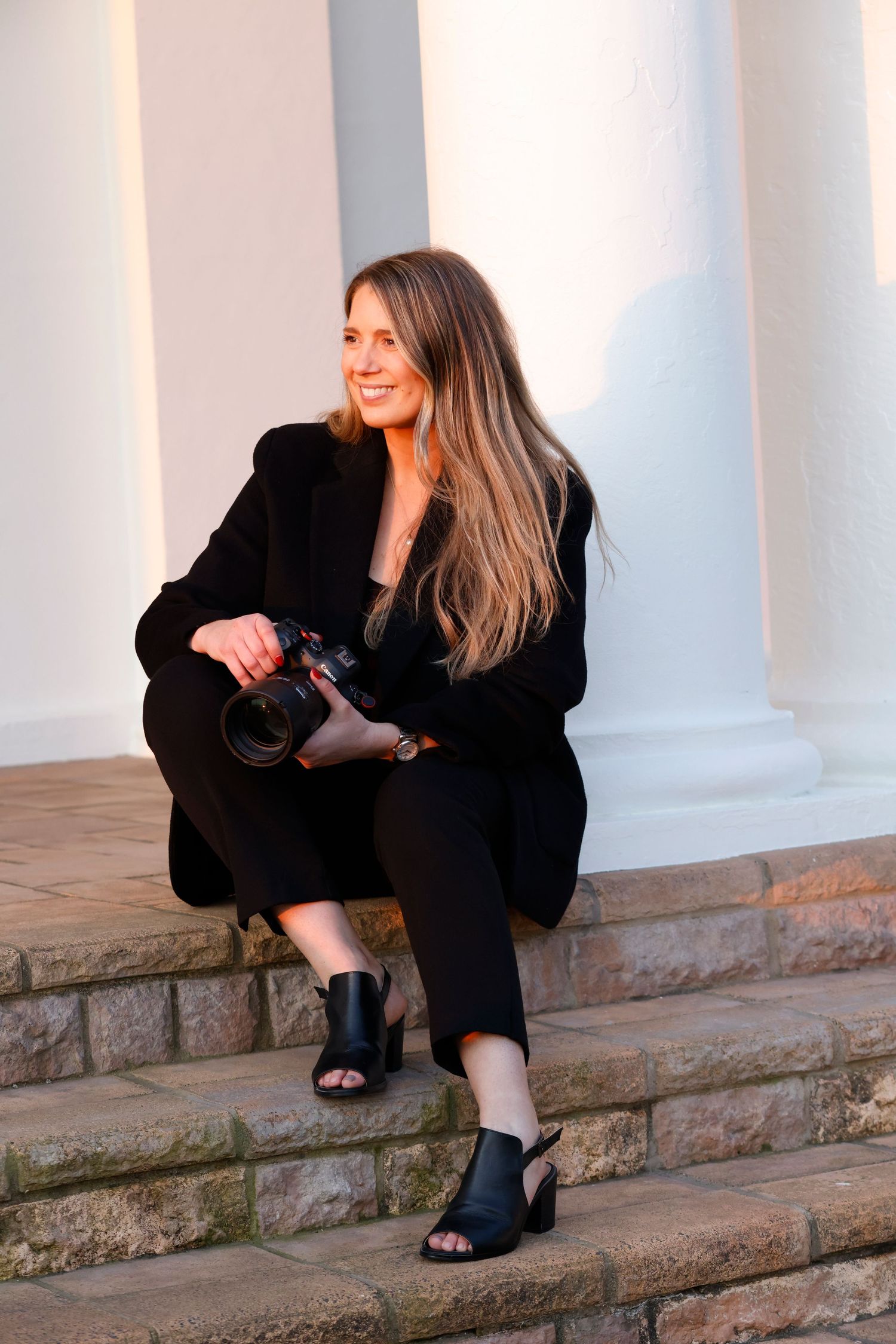 A photographer in black attire crouches on stone steps near white columns while taking pictures with a camera.