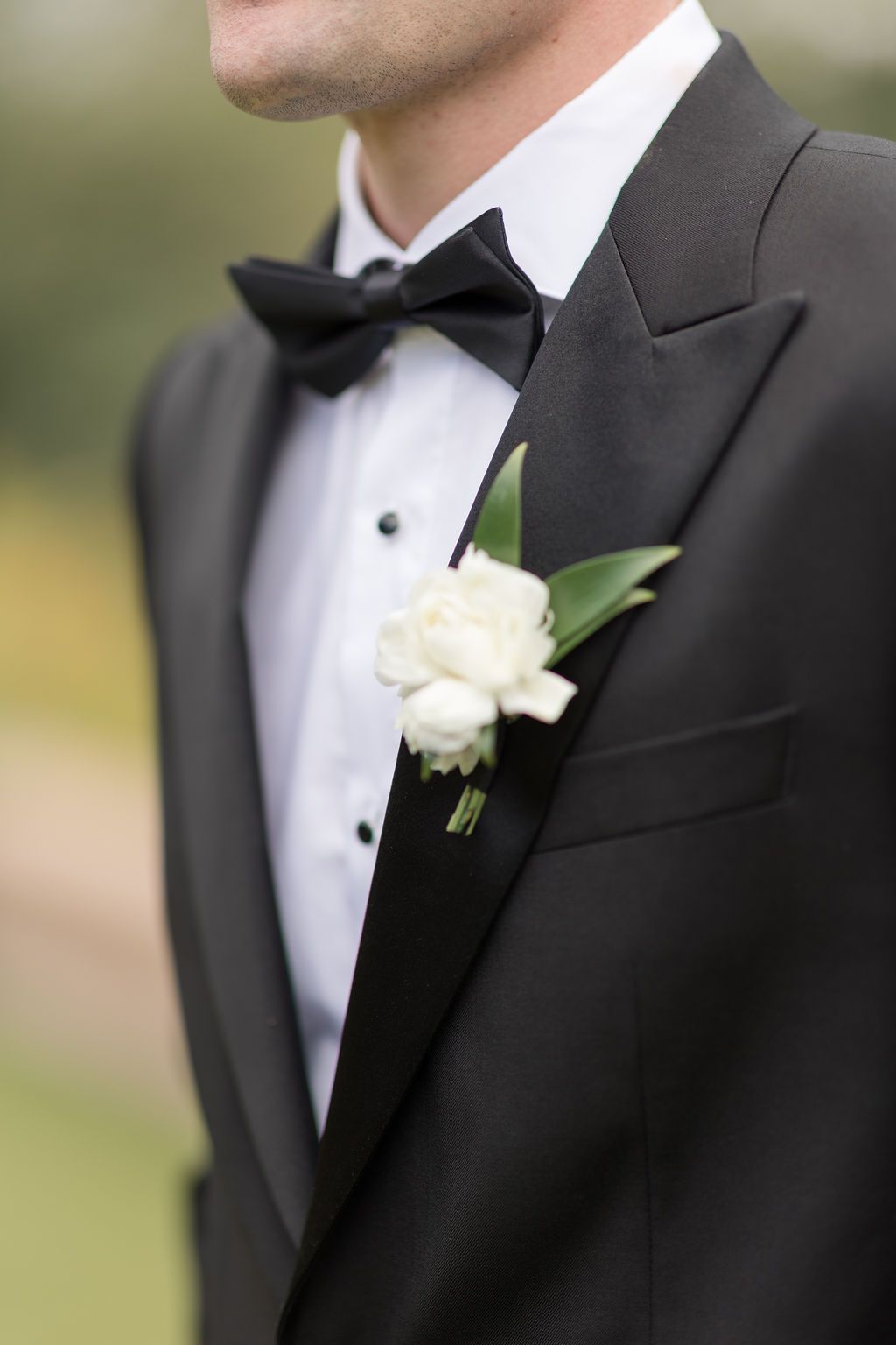 Groom in classic black tuxedo and bowtie smiles while standing near windows at wedding venue.