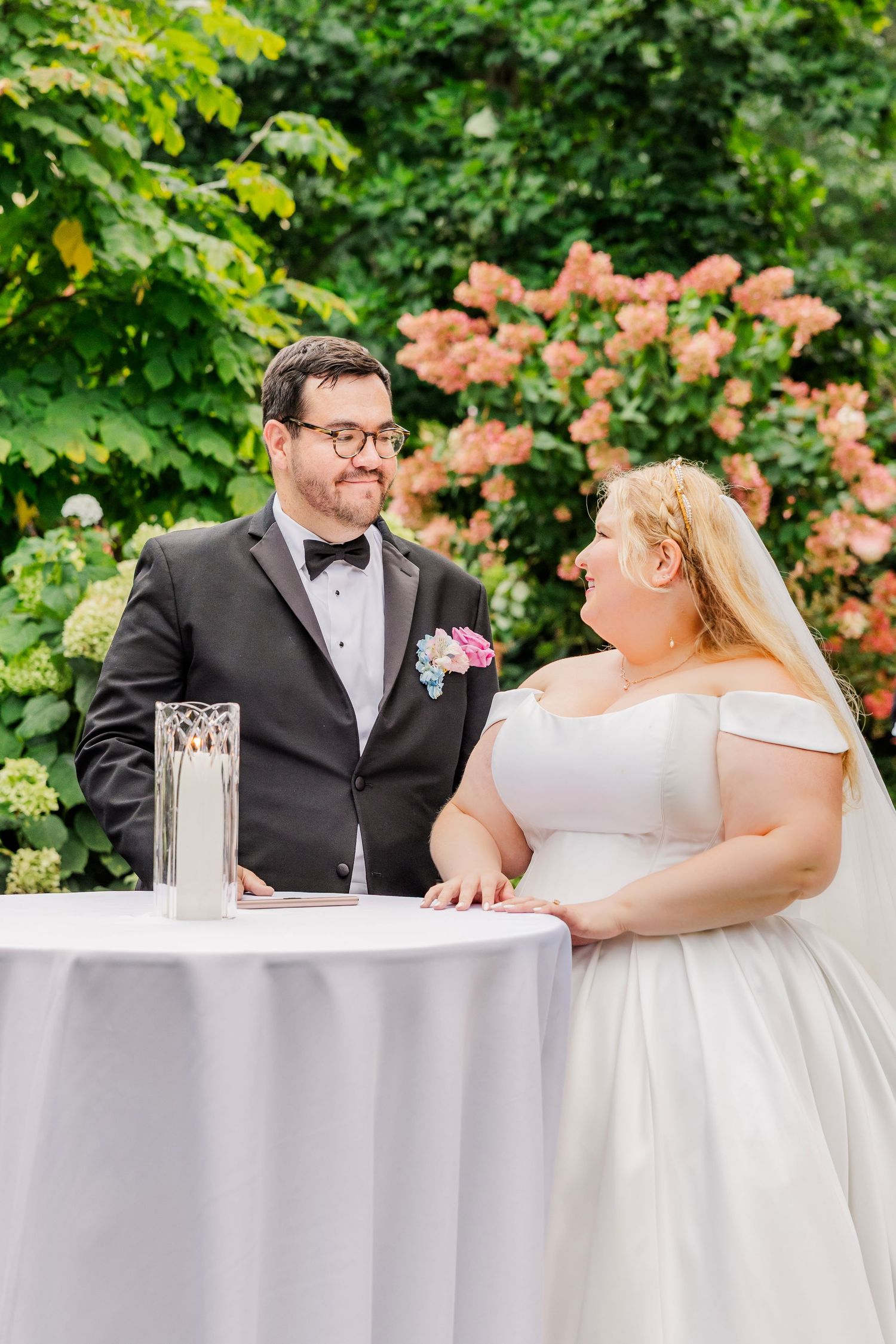Newlywed couple shares a romantic moment at an outdoor reception with coral flowers and greenery in the background.