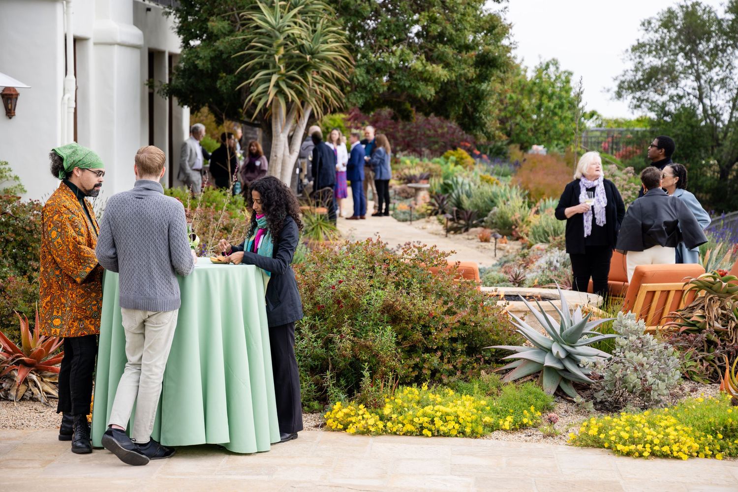 Guests mingling at an elegant outdoor garden party with desert landscaping and green tablecloths.