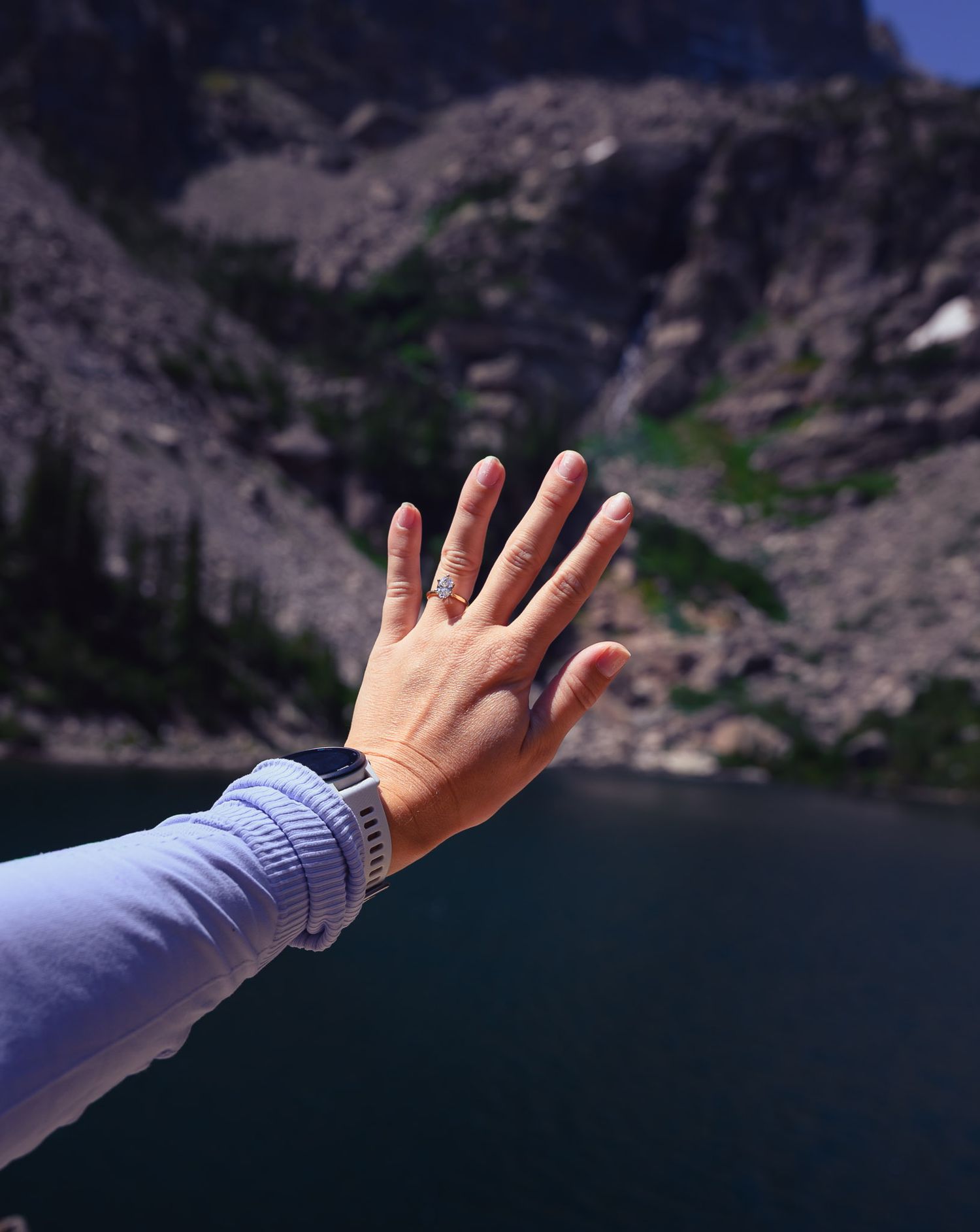 Close-up of a hand wearing an engagement ring with a scenic mountain lake in the background.